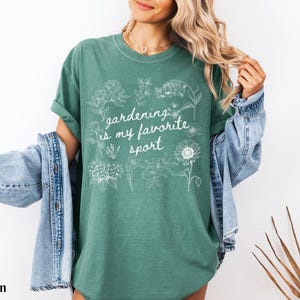May include: Light green t-shirt with the text "gardening is my favorite sport" in white script. The shirt features white floral illustrations. A denim jacket is draped over the shoulders.