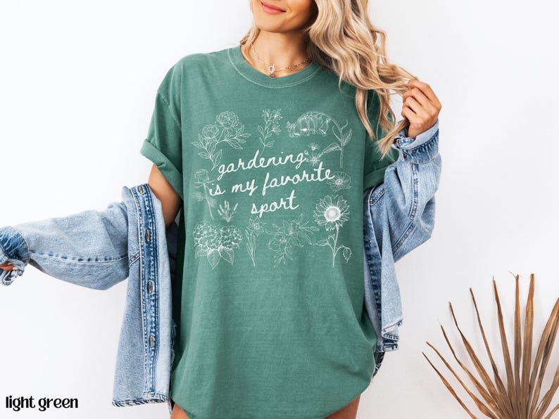 May include: Light green t-shirt with the text "gardening is my favorite sport" in white script. The shirt features white floral illustrations. A denim jacket is draped over the shoulders.