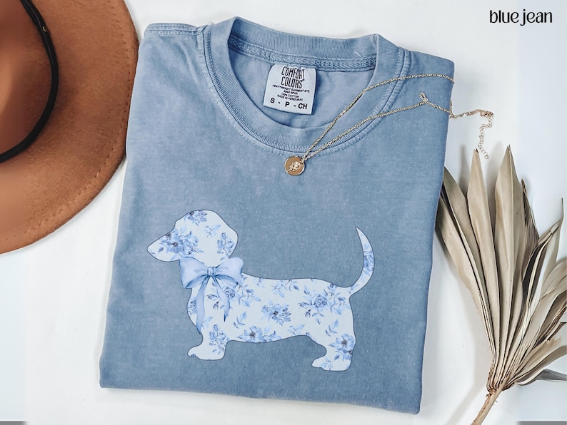 May include: A blue jean colored t-shirt with a floral dachshund design. The dachshund has a blue bow and is covered in a blue floral pattern. A gold necklace with a pendant is on the shirt. The shirt is folded on a white surface.