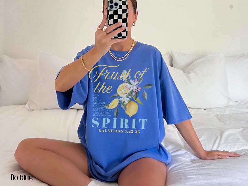 Op de afbeelding: Een blauw T-shirt met de tekst "Fruit of the Spirit" en een bloemen- en citroenafbeelding. De tekst "Liefde, Vrede, Geduld, Vriendelijkheid, Goedheid, Trouw, Zachtmoedigheid, Zelfbeheersing" staat ook op het shirt. De tekst "Galaten 5:22-23" staat eronder.