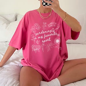 May include: A bright pink t-shirt with the text "gardening is my favorite sport" in white, surrounded by floral illustrations. The shirt is a loose fit and appears to be made of soft material.