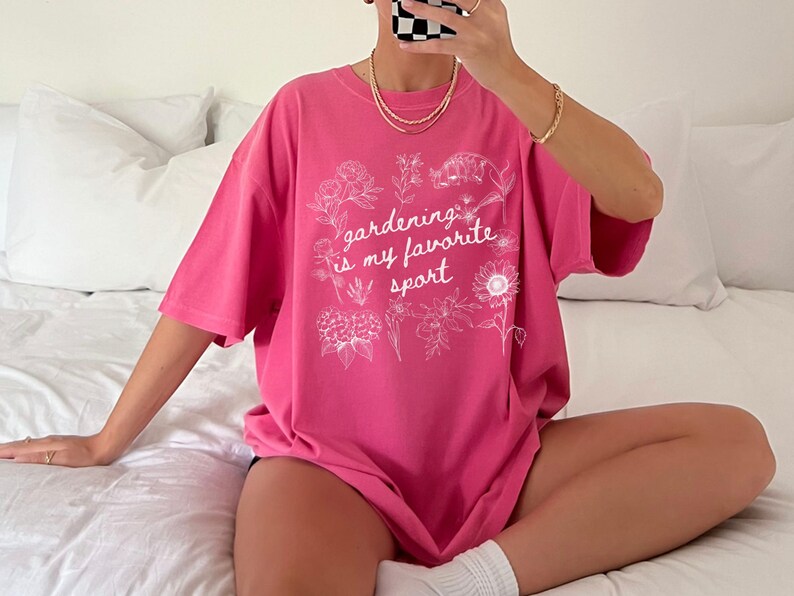 May include: A bright pink t-shirt with the text "gardening is my favorite sport" in white, surrounded by floral illustrations. The shirt is a loose fit and appears to be made of soft material.