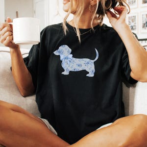 May include: Black t-shirt featuring a light blue dachshund dog design with a floral pattern and a bow. The dog is the focal point of the shirt. The model is holding a white mug.