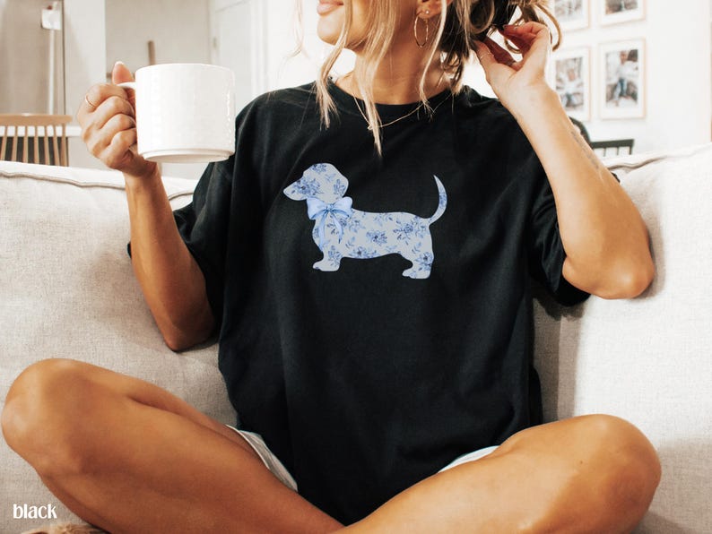 May include: Black t-shirt featuring a light blue dachshund dog design with a floral pattern and a bow. The dog is the focal point of the shirt. The model is holding a white mug.