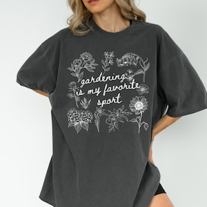 May include: Dark gray oversized t-shirt with the phrase "gardening is my favorite sport" in white script and floral illustrations. The shirt has short sleeves and a relaxed fit, perfect for casual wear.