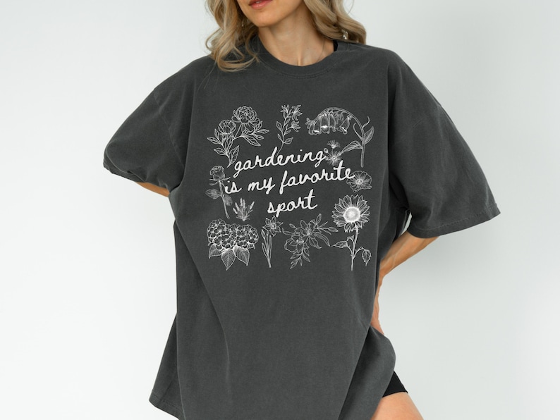 May include: Dark gray oversized t-shirt with the phrase "gardening is my favorite sport" in white script and floral illustrations. The shirt has short sleeves and a relaxed fit, perfect for casual wear.