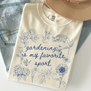 May include: Cream-colored t-shirt with blue floral illustrations and the text "gardening is my favorite sport." The shirt is laid on top of light blue jeans and a tan hat. A silver necklace is visible.