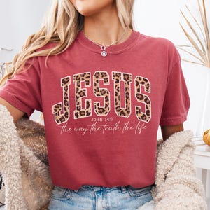 May include: Crimson-colored t-shirt with the word "JESUS" in large, leopard-print letters. Below the word "JESUS" is the text "John 14:6" and "the way the truth the life" in a white script font. The shirt has short sleeves.