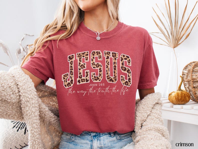 May include: Crimson-colored t-shirt with the word "JESUS" in large, leopard-print letters. Below the word "JESUS" is the text "John 14:6" and "the way the truth the life" in a white script font. The shirt has short sleeves.