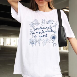 May include: White t-shirt with a blue floral design and the text "gardening is my favorite sport." The design includes various flowers and foliage. The shirt is a casual, comfortable style.