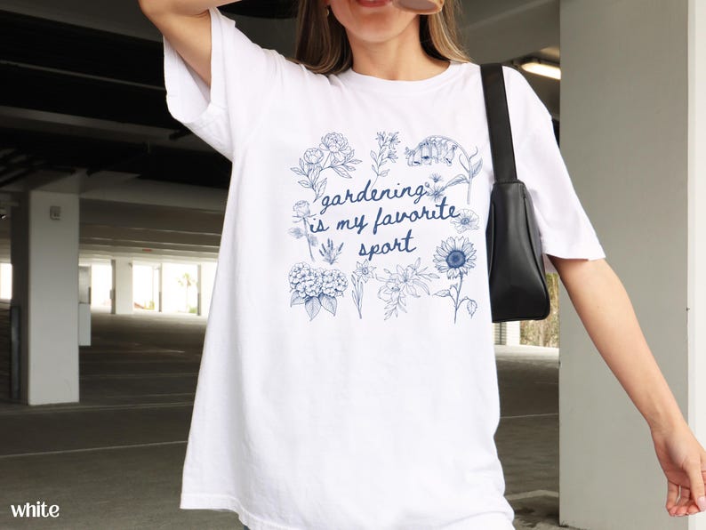 May include: White t-shirt with a blue floral design and the text "gardening is my favorite sport." The design includes various flowers and foliage. The shirt is a casual, comfortable style.