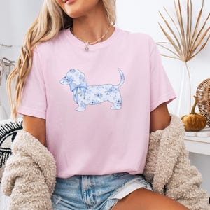 May include: A light pink t-shirt featuring a watercolor illustration of a dachshund dog with a blue floral pattern and a matching bow. The dog is the focal point of the shirt, which is made of soft material.