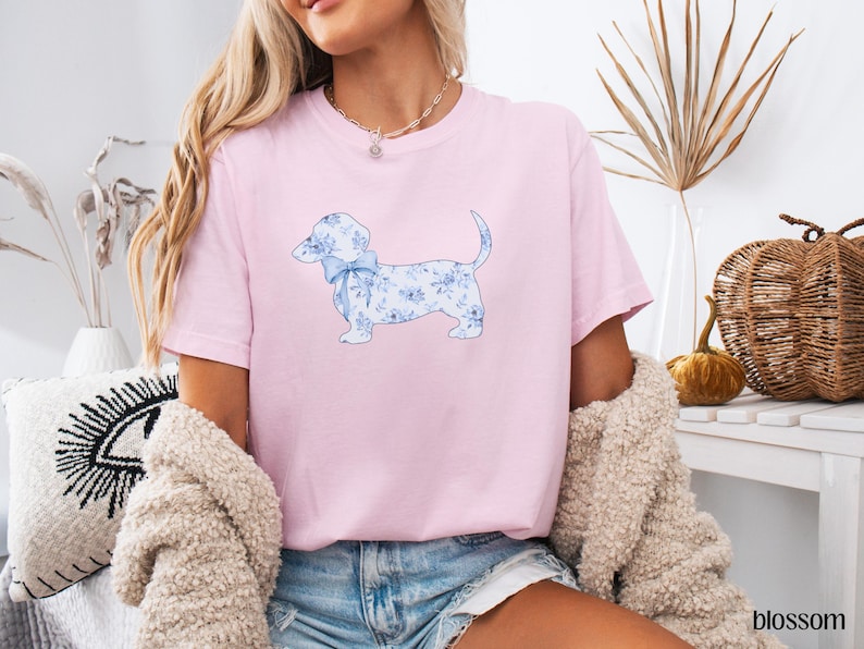 May include: A light pink t-shirt featuring a watercolor illustration of a dachshund dog with a blue floral pattern and a matching bow. The dog is the focal point of the shirt, which is made of soft material.