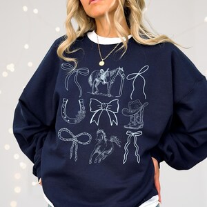 May include: Navy blue sweatshirt featuring white line drawings of equestrian and Western-themed elements, including a horse, cowboy hat, horseshoe, and bows. The word "navy" is printed at the bottom.