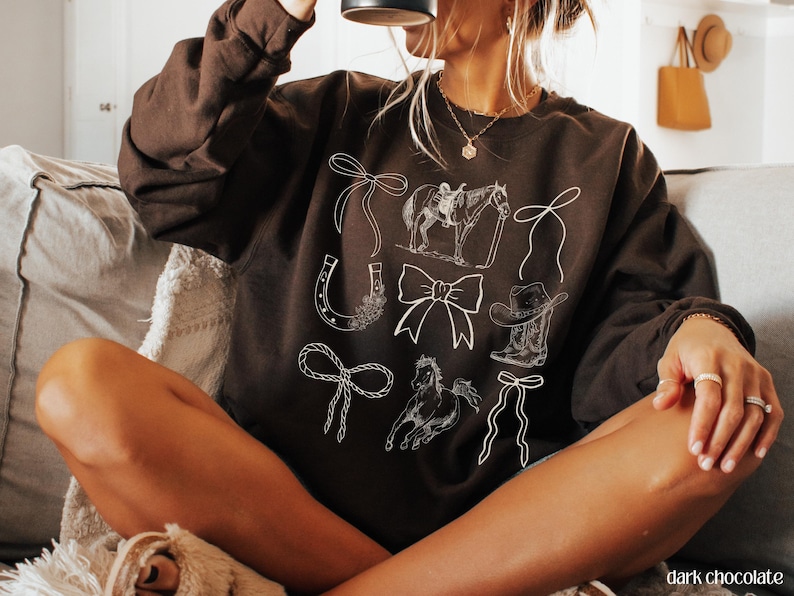 May include: Dark chocolate-colored sweatshirt featuring white line drawings of cowboy-themed elements. The design includes a horse and rider, bows, a horseshoe, a cowboy boot, and a cowboy hat. The text "dark chocolate" is visible at the bottom right.