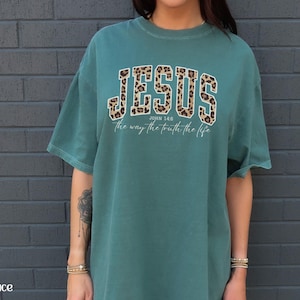 May include: Teal-colored t-shirt with the word "JESUS" in a leopard print design across the chest. Below the word "JESUS" is the text "John 14:6 the way the truth the life" in a white font.