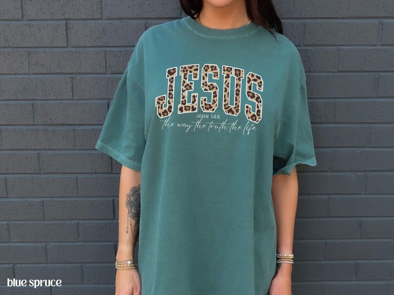 May include: Teal-colored t-shirt with the word "JESUS" in a leopard print design across the chest. Below the word "JESUS" is the text "John 14:6 the way the truth the life" in a white font.