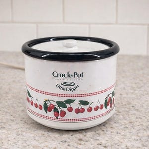 May include: White Crock-Pot Little Dipper slow cooker with a black rim and lid. The slow cooker has a cherry design around the base and the Crock-Pot logo. The slow cooker is on a light-coloured countertop.