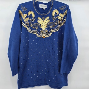 May include: A blue sweater with gold sequin and bead embellishments. The sweater features a round neckline and long sleeves. The front of the sweater is adorned with a large floral design made of gold sequins and beads. The brand name "VICTORIA HARBOUR" is visible on the label.