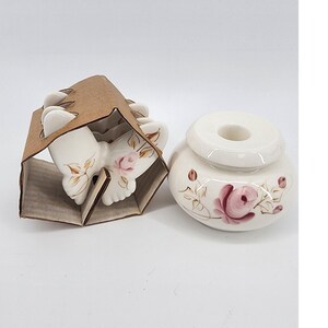 Può includere: Un set di sottobicchieri in ceramica bianca a forma di farfalla con accenti di rose rosa e dettagli di foglie dorate, esposti in un supporto di cartone marrone, accanto a un portacandele abbinato con un motivo a rosa rosa.