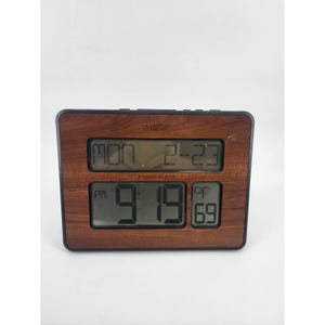 May include: A rectangular digital atomic clock with a wood-grain finish. The display shows the day, date, time, and temperature in Fahrenheit. The clock has the text "LA CROSSE TECHNOLOGY" at the top.