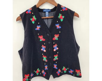 Op de afbeelding: Een zwart fluwelen vest met een V-hals en een knoopsluiting aan de voorzijde. Het kledingstuk is versierd met geborduurde kleurrijke kerstsokmotieven in rood, groen en blauw, geaccentueerd met kleine gouden kraaltjes. Een Karen Scott label is zichtbaar.