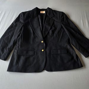 Puede incluir: Un blazer negro con dos botones dorados y dos bolsillos delanteros. El blazer tiene una solapa con muescas clásica y mangas largas. La prenda está hecha de una tela oscura y está tendida sobre una superficie gris.