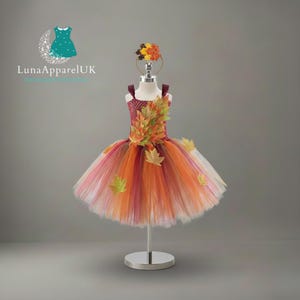 Puede incluir: Vestido tutú con temática otoñal y diadema a juego. El vestido presenta un corpiño con estampado de lunares burdeos y una falda en capas en tonos naranja, rojo y blanco. Adornos de hojas decorativas en el corpiño y la falda.