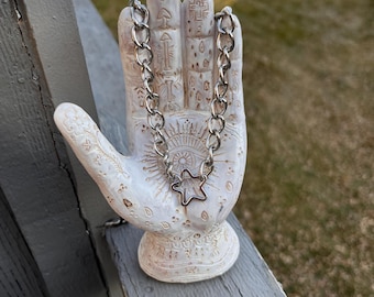 May include: A silver-colored chain necklace with a star pendant rests on a white ceramic hand sculpture. The sculpture is detailed with etched designs and sits on a weathered wooden surface, with a blurred outdoor background.