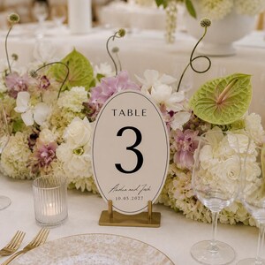 May include: An oval-shaped table number sign featuring the text TABLE 3, Amelia and Josh, and the date 10.05.2027. The sign is cream-colored with a thin black border, held upright by a rectangular gold-toned metal stand on a white tablecloth.