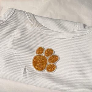 Pode incluir: Body branco canelado com um design de pata dourada com contas. A pata está centrada no peito. O body está dobrado sobre uma superfície branca.