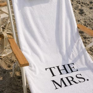 May include: White beach towel with the text "THE MRS." in black, draped over a white beach chair with wooden armrests. A small beige pillow rests at the top of the chair. The scene is set on a sandy beach.