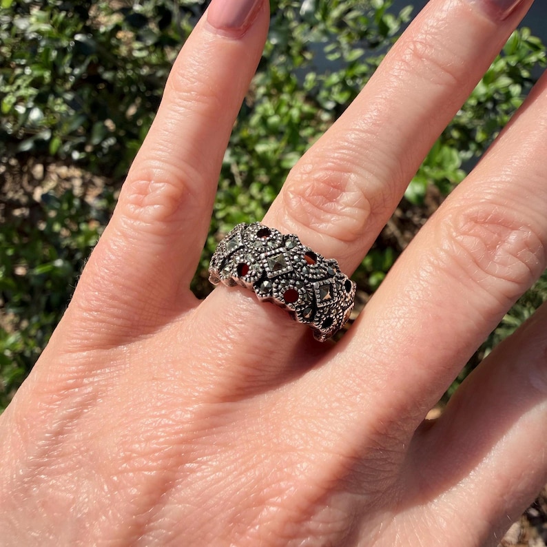 May include: A silver-toned ring with a detailed pattern of small, square-shaped marcasite stones and interspersed red gemstones. The ring is worn on a finger, showcasing its intricate design and craftsmanship. The ring's design is a continuous band.