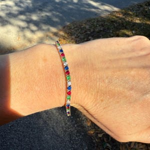 Pode incluir: Uma pulseira dourada com uma fileira de pedras preciosas coloridas. As pedras são vermelhas, azuis, verdes e brancas. A pulseira é usada no pulso.