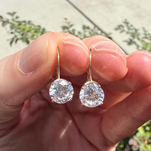 May include: A pair of gold leverback earrings featuring round, clear, faceted stones set in a prong setting. The earrings have a polished gold finish and a classic circular design that catches the light, showcasing the brilliance of the stones.