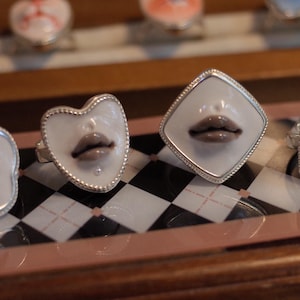 May include: Four silver rings featuring white ceramic faces with sculpted, glossy brown lips. The rings have distinct shapes including a flower, a heart, a diamond, and a square, all set in metallic frames with a beaded edge detail.