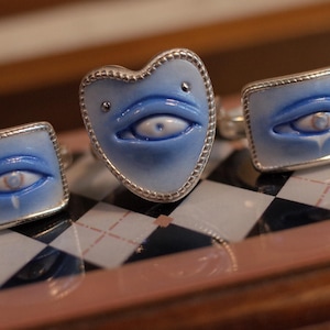 May include: A set of jewelry consisting of one heart-shaped ring and two rectangular cufflinks. Each piece features a light blue, smooth surface with a sculpted, realistic human eye in the center, all encased in a thin, silver-toned metal border.