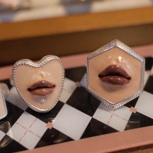 May include: Two silver-toned rings featuring a sculpted beige face detail with glossy brown lips. One ring has a heart-shaped frame, while the other is hexagonal. Both pieces display a realistic, three-dimensional lip design set within a metallic border.