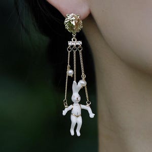 May include: Gold-toned earrings featuring a lion's head stud, dangling chains, and a white rabbit figurine. The earrings include small pearls and the text "Flame Essence x YIHE". The design is intricate and whimsical, suitable for a unique jewelry collection.