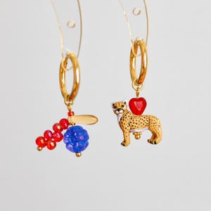 May include: Gold hoop earrings with unique charms. One earring features a cluster of red beads and a blue bead, the other has a cheetah charm and a red heart. The earrings hang against a white background.
