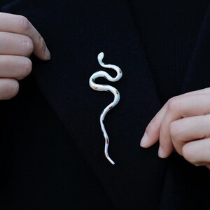 May include: A white snake-shaped brooch with gold accents is pinned to a black garment. The brooch has a curved design and is made of a glossy material. The text "Flame Essence x YIHE" is at the bottom.