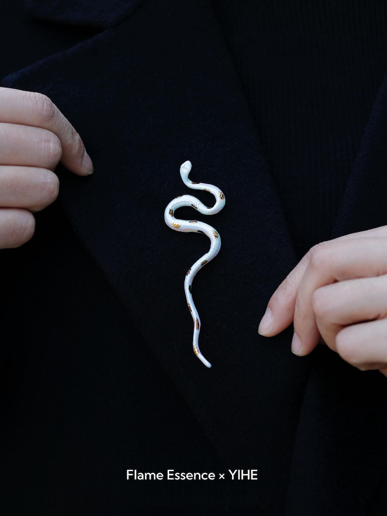 May include: A white snake-shaped brooch with gold accents is pinned to a black garment. The brooch has a curved design and is made of a glossy material. The text "Flame Essence x YIHE" is at the bottom.