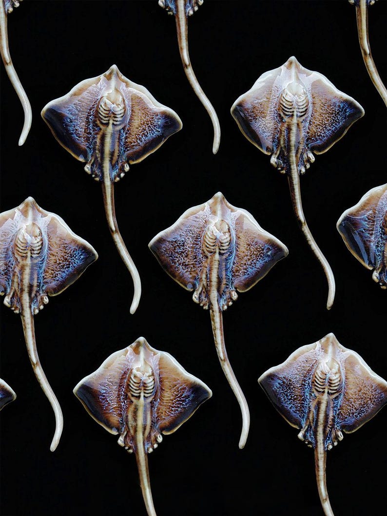 K&ouml;nnte beinhalten: Ein sich wiederholendes Muster aus Keramik-Stechrochen-Skulpturen. Jede Skulptur hat einen braunen und blau get&ouml;nten K&ouml;rper mit einem wei&szlig;en Schwanz. Die Rochen sind vor einem schwarzen Hintergrund angeordnet.