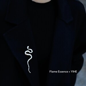 May include: A silver-colored snake brooch pinned to a black coat. The brooch is a stylized representation of a snake with a curved body. The text "Flame Essence x YIHE" is visible in the lower right corner.