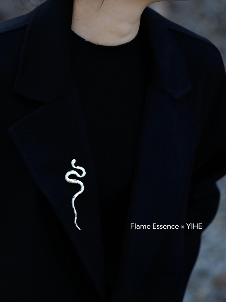 May include: A silver-colored snake brooch pinned to a black coat. The brooch is a stylized representation of a snake with a curved body. The text "Flame Essence x YIHE" is visible in the lower right corner.