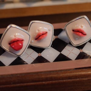 May include: Three square-shaped cufflinks featuring a pale, skin-toned base with a raised, realistic depiction of human lips in a vibrant red color. One cufflink shows a tongue sticking out, while the others show closed or slightly parted lips.
