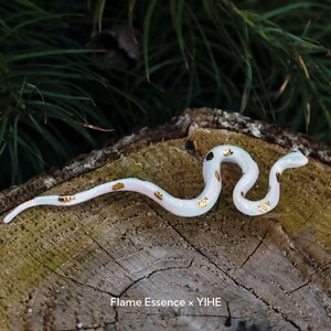 May include: A white and gold decorative snake figurine with a pearlescent finish. The snake has gold accents and is posed on a wooden surface. The text "Flame Essence x YIHE" is visible at the bottom.