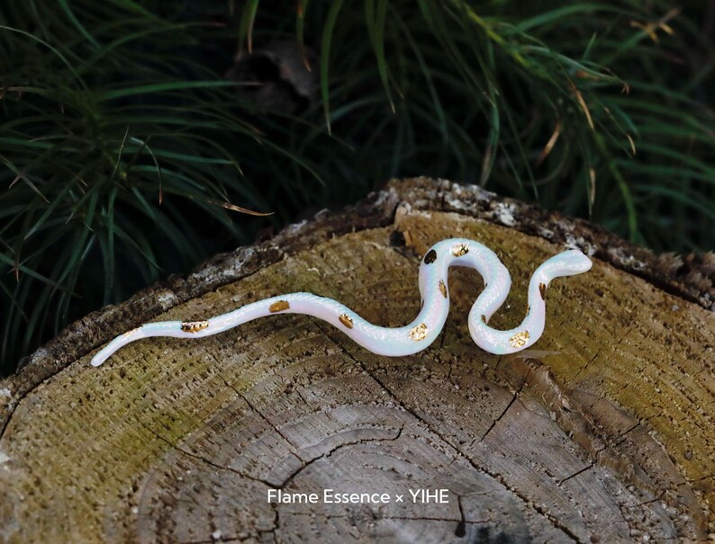 May include: A white and gold decorative snake figurine with a pearlescent finish. The snake has gold accents and is posed on a wooden surface. The text "Flame Essence x YIHE" is visible at the bottom.