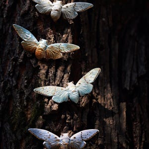 Puede incluir: Cuatro esculturas decorativas de cigarras con las alas abiertas están fijadas a un tronco de árbol. Las cigarras tienen alas azules y blancas con detalles en marrón y dorado. El texto "Flame Essence x YIHE" está en la parte inferior.