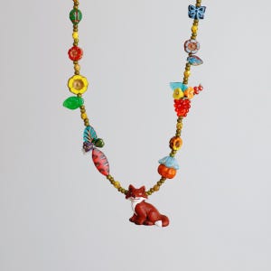 May include: A colorful beaded necklace featuring a fox pendant. The necklace includes various beads and charms, such as flowers, leaves, and a butterfly. The fox pendant is brown and white. The necklace is a whimsical accessory.